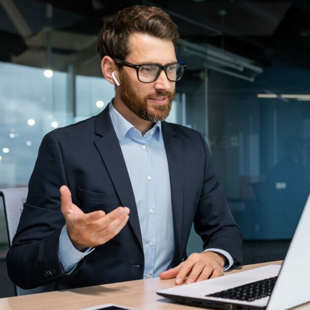 Ein Mann im Anzug und Kopfhörern in den Ohren sitzt an einem Schreibtisch vor einem aufgeklappten Laptop und gestikuliert mit der Hand in einem Raum mit Glaswand.