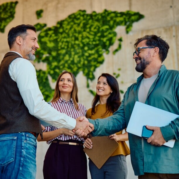 Zwei Männer mit Laptops im Arm geben sich lächelnd die Hand, daneben stehen zwei Frauen vor einer hohen Wand mit abstrakter, grüner Weltkarte.