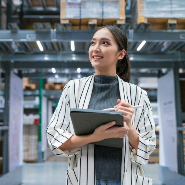 Eine lächelnde Frau steht mit Tablet und Stift in den Händen in einer Lagerhalle.