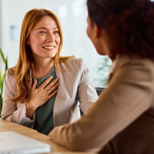 ine Frau im Blazer hat eine Hand auf die Brust gelegt und lächelt eine weitere Frau an, beide sitzen an einem Tisch, auf dem ein Tablet und ein Wasserglas stehen.