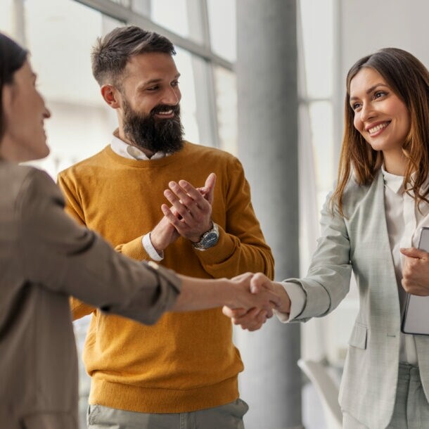 Vier Personen stehen beieinander, zwei Frauen geben sich lächelnd die Hand, während zwei Männer in die Hände klatschen.