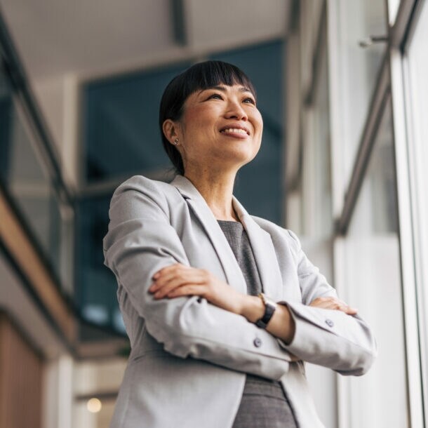 Porträt einer lächelnden Frau im Blazer mit verschränkten Armen in einem Bürogebäude mit viel Glas.