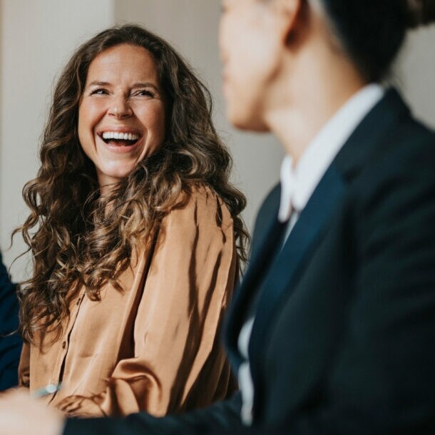 Eine lachende Frau sitzt vor einem Laptop an einem Tisch zwischen zwei weiteren Personen in Business-Kleidung während eines Meetings.