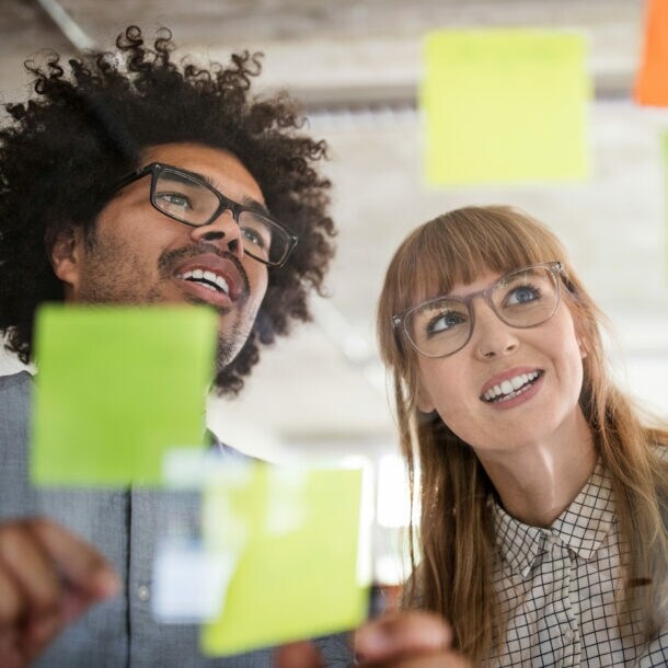 Nahaufnahme von Mann und Frau hinter einer Glasscheibe mit bunten Klebezetteln in einem Büro.