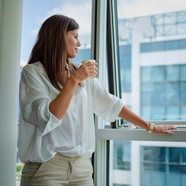 Eine Frau in weißer Bluse steht mit einem Kaffeebecher in der Hand an einem halbgeöffneten Fenster und blickt hinaus auf ein gläsernes Gebäude.