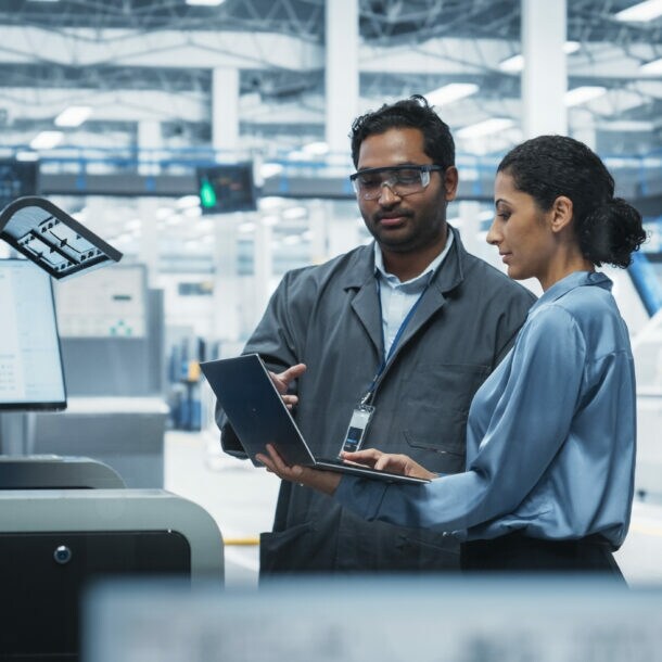 Ein Mann und eine Frau stehen an einer digitalen Schaltzentrale einer Elektrofabrik und schauen gemeinsam auf ein Laptop.