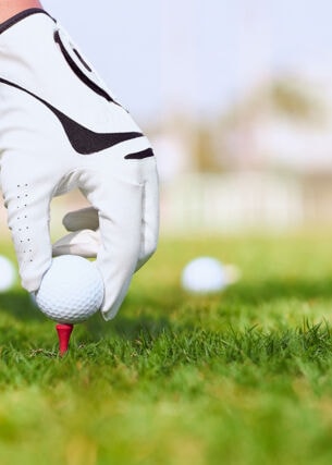 Detailaufnahme von Hand mit Handschuh, die Golfball auf Rasen platziert, daneben Golfschläger