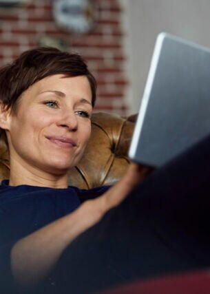 Eine Frau auf einer Ledercouch schaut zufrieden in ihr Tablet