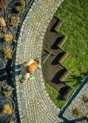 Luftaufnahme einer Person, die auf einem gepflasterten Weg stehend eine Fläche mit Rollrasen in einem Garten bearbeitet