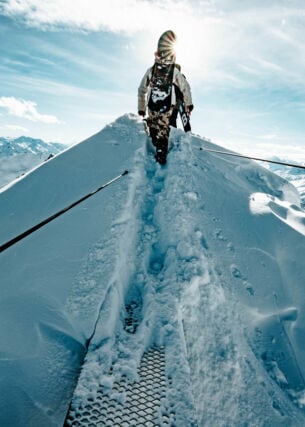 Ein:e Wintersportler:in mit Snowboard auf dem Rücken geht über einen schneebedeckten Berggipfel.