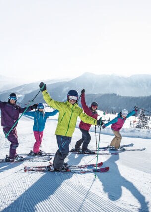 Fünf Skifahrende in bunter Kleidung winken in die Kamera auf einer Piste vor winterlichem Bergpanorama