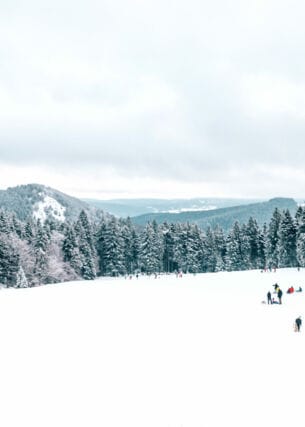 Eine schneebedeckte Freifläche mit Menschen, im Hintergrund Tannenwälder und Bergketten.