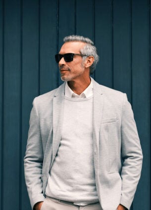 Mann vor einer blauen Holzwand mit weißem Pullover und hellem Anzug mit Sonnenbrille