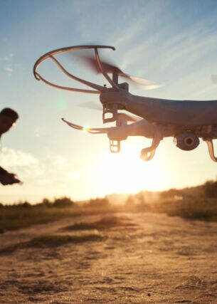 Eine Drohne mit Kamera an der Unterseite fliegt im Tiefflug über ein Feld im Sonnenuntergang, im Hintergrund der Pilot als Silhouette