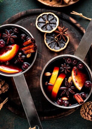 Heißes Getränk gekocht in zwei Topf mit Gewürzen, Orangen und Preiselbeeren, umrandet von Tannenzapfen und Tannenzweigen