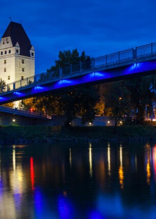 Das beleuchtete Neue Schloss in Ingolstadt bei Nacht, im Vordergrund führt eine Brücke über die Donau