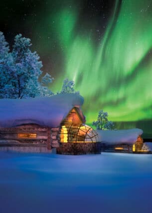 Zwei erleuchtete und schneebedeckte Blockhütten mit Glasanbau in Winterlandschaft mit grünem Polarlicht am nächtlichen Himmel