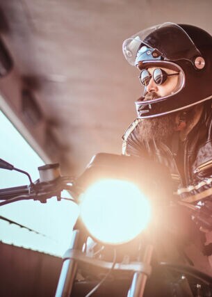 Ein junger, bärtiger Mann in Lederjacke lehnt lässig mit Helm und Sonnenbrille auf einem Motorrad und blickt aus einem offenen Betonbauwerk in die Ferne