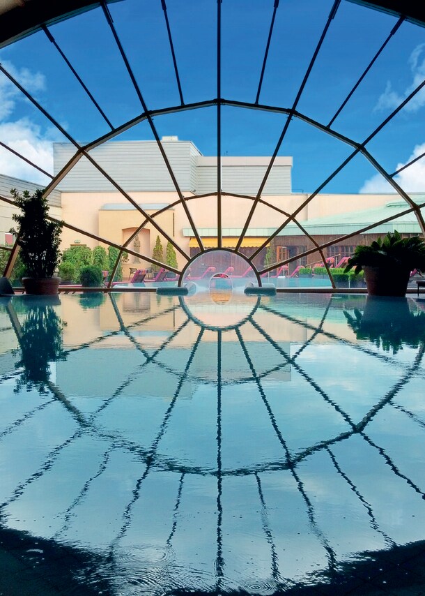 Ein großer Innenpool führt durch ein großes, rundes Panoramafenster in den Außenbereich mit Terrasse