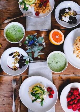 Topshot von unterschiedlichen vegetarischen Speisen auf weißem Geschirr auf Holztisch, umgeben von Besteck, Servietten und Pflanzen