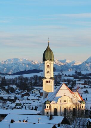 Schneebedeckter Ort mit weißer Kirche vor Alpenpanorama in der Dämmerung
