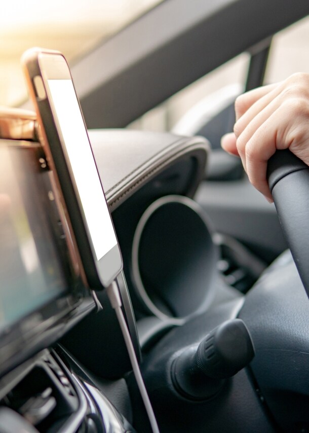 Ein Smartphone in einer Halterung am Armaturenbrett eines Autos, daneben zwei Hände an einem Lenkrad