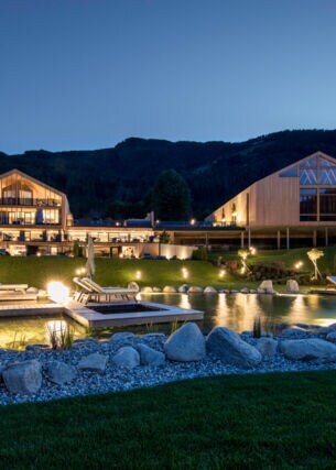 Eine luxuriöse, beleuchtete Hotelanlage aus Holzgebäuden mit Landschaftsgarten und Badeteich vor Bergpanorama bei Nacht