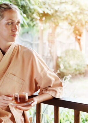Eine junge Frau sitzt im Morgenmantel auf einer Terrasse und hält eine Tasse Tee in ihren Händen