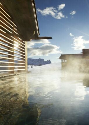 Ein Pool in der strahlenden Sonne, daneben Holzhäuser und Berge im Hintergrund