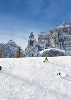 Panorama einer Skipiste mit drei Skifahrern vor markanten Felsformationen eines Bergmassivs
