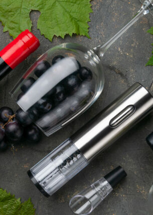 Aufsicht von elektrischem Metall-Korkenzieher, Weinbbelüfter und Vakuumstopfen neben Weinflaschen, Gläsern und Trauben auf grauem Betonhintergrund