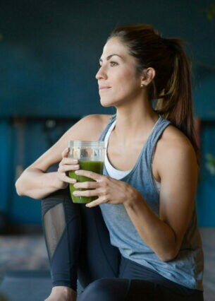 Eine junge Frau sitzt in Sportkleidung mit einem grünen Saft in ihren Händen auf einer Yogamatte in einem Raum mit Grünpflanzen.