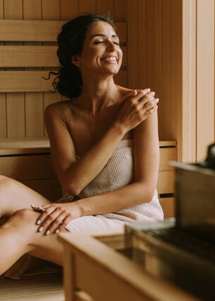 Eine lachende Frau sitzt eingewickelt in ein handtuch in einer Holzsauna.