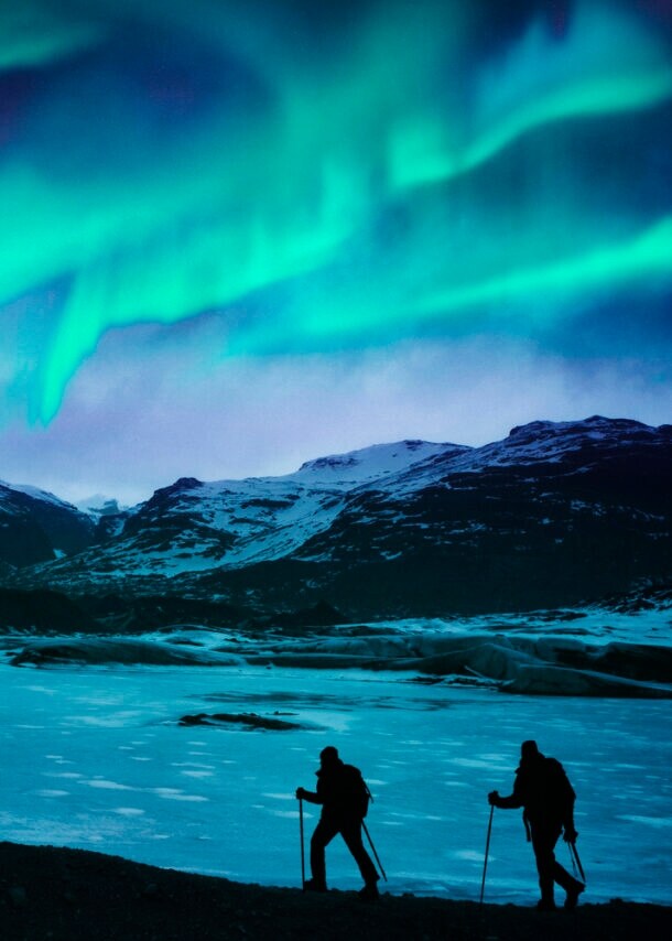 Zwei Wanderer vor einer Bergkulisse mit Polarlichtern.
