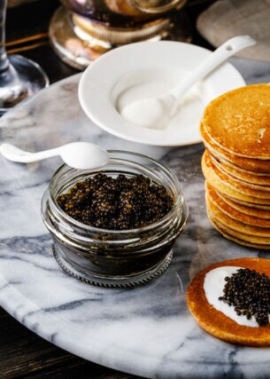 Kaviar im Glas, ein Stapel Blinis und Löffel auf einem Marmortisch, daneben Sektkorken und -gläser