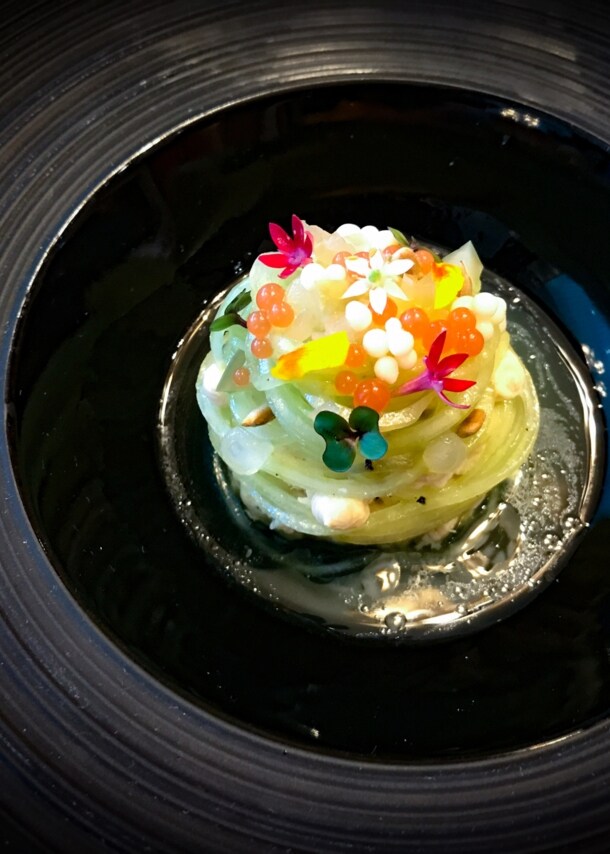 Nahaufnahme eines Gourmet-Gerichtes mit grünen, zusammengerollten Nudeln, dekoriert mit Kaviar und Blüten und angerichtet auf einem schwarzen Teller