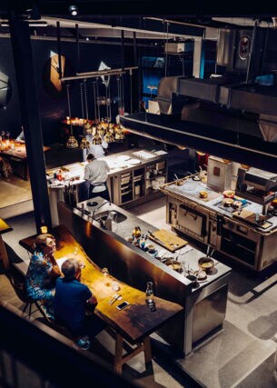 Blick in ein Restaurant mit Holztischen und mittiger Kücheninsel