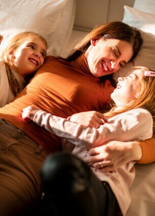 Eine Frau kuschelt lachend mit ihren zwei Töchtern im Bett