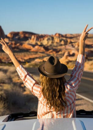 Eine junge Frau ragt mit dem Oberkörper aus dem Schiebedach eines Autos auf einer Straße in einer Wüstenlandschaft und winkt spontan mit einem Hut in die Luft