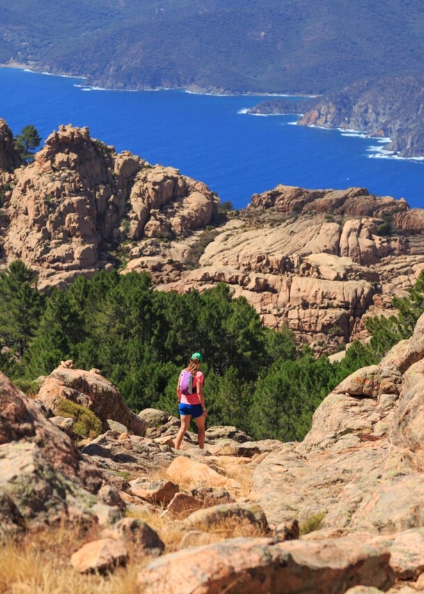 Eine Frau wandert über felsiges Gebiet, im Hintergrund ist das Meer zu sehen.