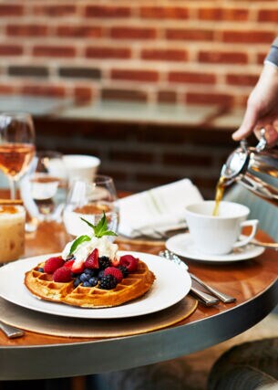 Eine Tasse Kaffee wird neben einem Teller mit Waffeln auf einem gedeckten Frühstückstisch eingegossen