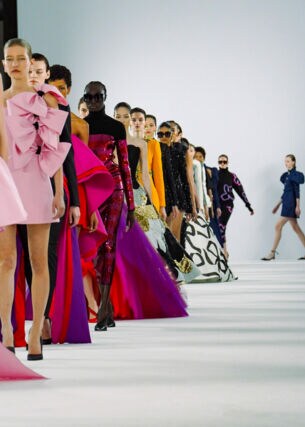 Eine Reihe weiblicher Models in bunten Abendkleidern vor Publikum auf einem Laufsteg