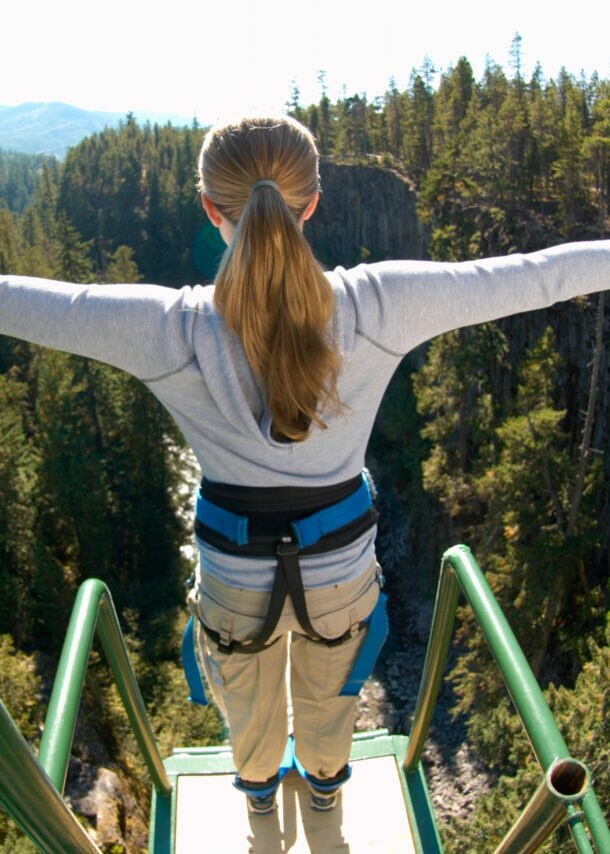 Eine Frau steht mit ausgebreiteten Armen gesichert an einem Bungee-Jumping-Gerüst und ist kurz davor runterzuspringen