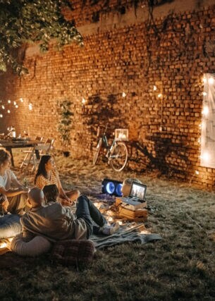 Eine Personengruppe sieht mit einem Mini-Beamer einen Film an einer Mauer im Garten.