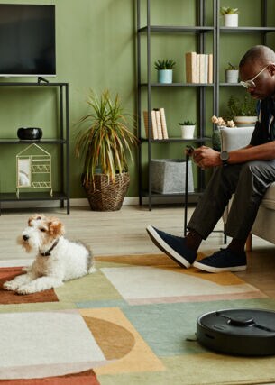 Ein Mann sitzt im Wohnzimmer auf dem Sofa und schaut auf sein Handy in der Hand. Vor ihm liegt ein Hund auf dem Teppich, daneben arbeitet ein Saugroboter.