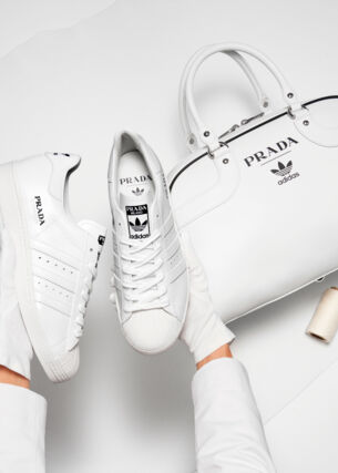 Weiße Turnschuhe neben einer Handtasche mit Logos der Marken adidas und Prada auf weißem Untergrund