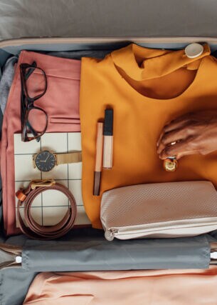 Eine Hand legt einen kleinen Gegenstand in einen geöffneten Koffer, in dem bereits Gegenstände wie eine Brille, eine Armbanduhr und Kleidung liegen