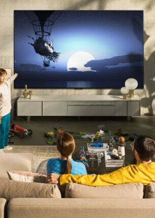Ein Mann und eine Frau sitzen auf einem beigen Sofa und schauen auf einen großen Fernseher, davor steht ein Junge und zeigt auf den Bildschirm