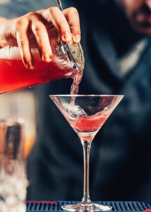 Nahaufnahme eines Cocktailglases, das von einem Barkeeper mit einer roten Flüssigkeit gefüllt wird