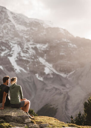 Zwei Personen sitzen im Nationalpark Stilfserjoch auf einem Stein und blicken auf ein Bergmassiv.